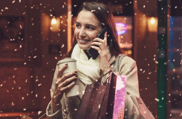 街を歩いてクリスマスショッピングをしている幸せな若い女性は 彼女は多くの袋を持っており 彼女のスマートフォンで電話を持っています — ストック写真