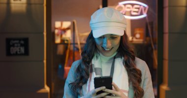 Şehir caddesinde yürüyen ve akıllı telefonuyla iletişim kuran mutlu, genç, modaya uygun bir kadın.