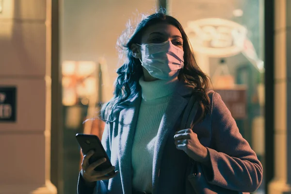 顔のマスクをした女性が通りを歩いていると 彼女は彼女のスマートフォン 背景にショップウィンドウを使用しています — ストック写真