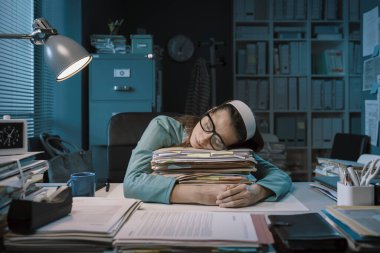 Exhausted office worker sleeping at desk and leaning on a pile of paperwork: overtime work and deadlines concept