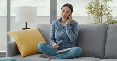 Gülümseyen kadın kanepede dinleniyor ve akıllı telefonuyla bir telefon görüşmesi yapıyor.