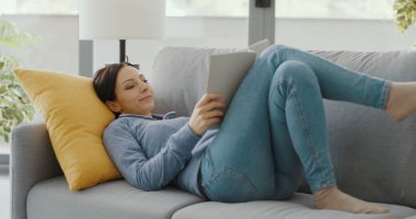 Evdeki kanepede dinlenen mutlu genç kadın, bir kitap okuyor.