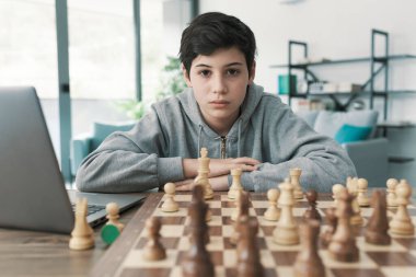 Çocuk evde satranç öğreniyor, dizüstü bilgisayar kullanıyor ve satranç tahtasına bakıyor.