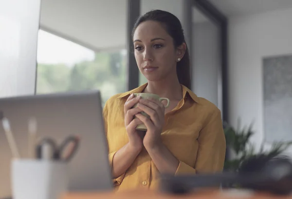 オフィスの机に座ってコーヒーを飲んでいる若い女性 彼女はノートパソコンの画面を見ている — ストック写真