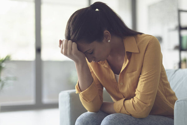 Depressed young woman sitting in the living room at home, she is feeling hopeless and sad