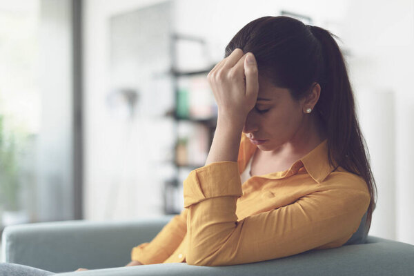 Depressed young woman sitting in the living room at home, she is feeling hopeless and sad