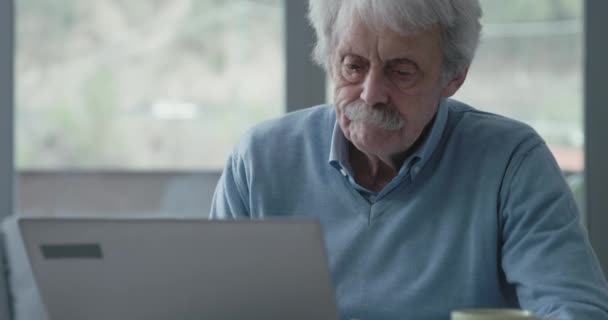 Senior man zit achter het bureau en gebruikt een laptop — Stockvideo