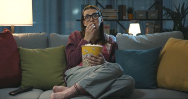 Televizyon seyreden ve patlamış mısır yiyen bir kadın evdeki kanepede oturmuş televizyon ekranına bakıyor.