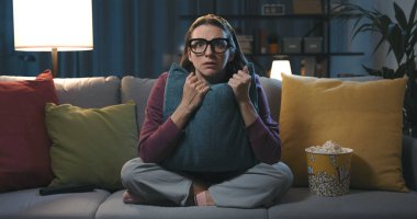 Kanepede oturup TV 'de korku filmi izleyen korkmuş bir kadın yastığa sarılıyor.