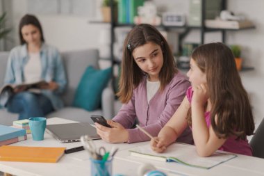 Happy girls sitting at desk at home and studying together, one of them is using a smartphone