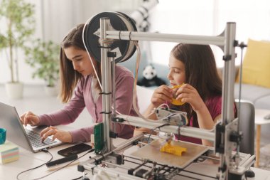 Young smart girls learning 3D printing at home, they are using a 3D printer and a laptop, science and education concept