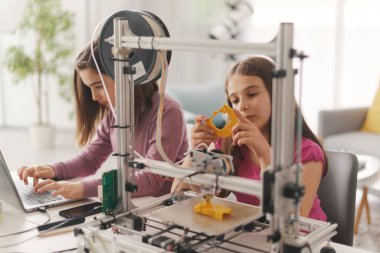 Young smart girls learning 3D printing at home, they are using a 3D printer and a laptop, science and education concept