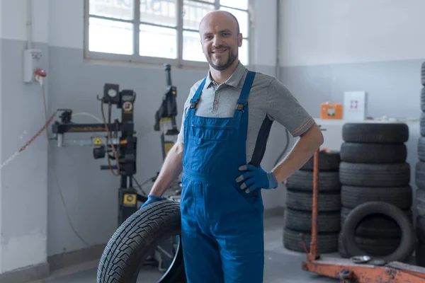 一位专业的微笑技师在汽车修理站摆姿势 他拿着一个轮胎 — 图库照片