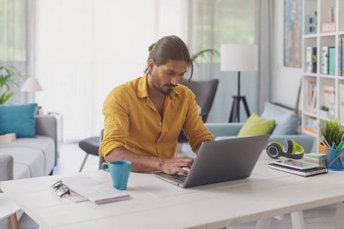 Genç adam masada oturuyor ve evde dizüstü bilgisayarıyla çalışıyor, uzaktan çalışma konsepti.
