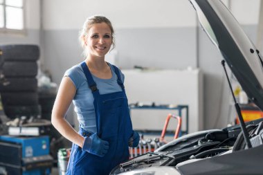 Oto tamirhanesinde arabanın yanında poz veren kendine güvenen kadın tamirci.