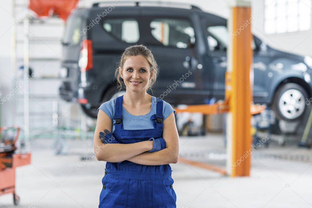Smiling young female mechanic posing in the car repair shop, she is looking at camera with arms crossed