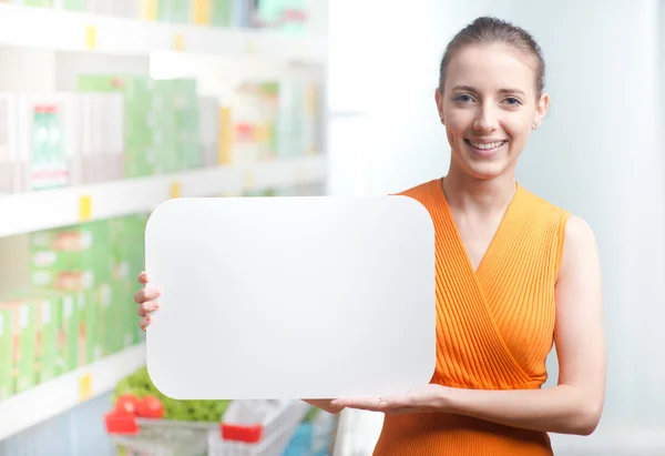 Smiling woman holding a white sign at supermarket — Stock Photo, Image