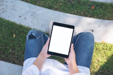 Açık havada otururken boş beyaz masaüstü ekran ile siyah tablet tableti tutan ve kullanan bir kadının en üst görünüm görüntüsü