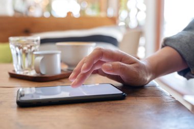 Kafedeki ahşap masada kahve fincanıyla akıllı bir telefon ekranını kullanan ve ona dokunan bir kadın eli.