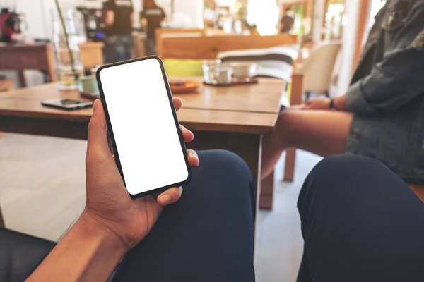 Mockup image of a man's hand holding black mobile phone with blank white screen with woman sitting in cafe