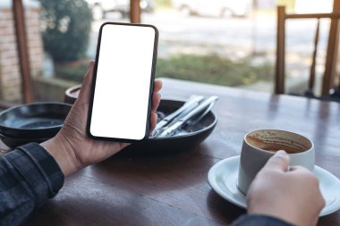 Kafede kahve içerken siyah cep telefonunu boş ekran ile tutan ellerin görüntüsü.
