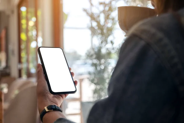 Mockup image of a woman holding black mobile phone with blank screen while drinking coffee in cafe