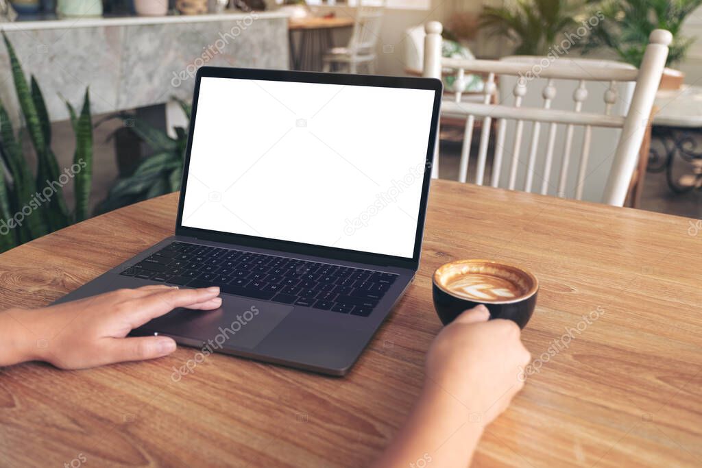 Mockup image of woman's hands typing and touching on laptop touchpad with blank white desktop screen while drinking coffee on wooden table in cafe
