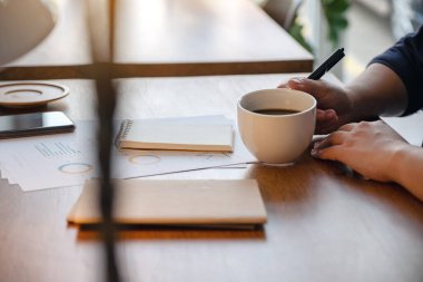 Ahşap masa üzerinde iş belgesi üzerinde çalışırken elinde bir fincan sıcak kahve tutan bir kadının yakın plan görüntüsü.