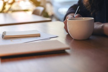 Ahşap masa üzerinde iş belgesi üzerinde çalışırken elinde bir fincan sıcak kahve tutan bir kadının yakın plan görüntüsü.