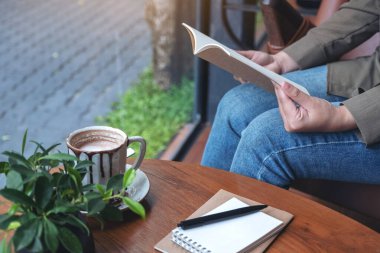 Kafe 'deki ahşap masada elinde defterler ve kahve fincanıyla kitap açan bir kadının yakın plan resmi. 