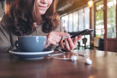 Kafedeki ahşap masada kulaklık ve kahve fincanı ile cep telefonu kullanan bir kadının yakın plan görüntüsü.