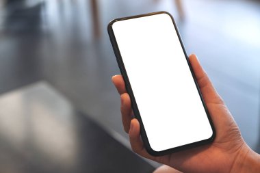 Kafedeki boş beyaz ekranlı siyah cep telefonlu bir kadının ellerinin görüntüsü.