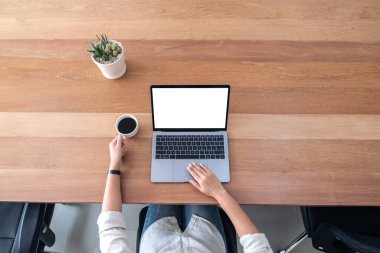 Ofisteki ahşap masada kahve içerken laptopun dokunmatik ekranını beyaz ekranla kullanan ve dokunan bir kadının üst görünüm görüntüsü