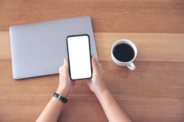 Üst görüntüde, elinde siyah cep telefonu olan, beyaz ekran, kahve fincanı ve dizüstü bilgisayarı olan bir kadının ahşap masa üzerinde duran resmi var.