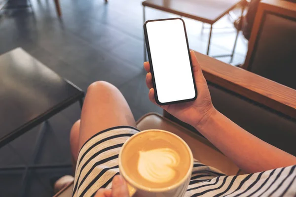 Mockup image of a woman holding black mobile phone with blank screen while drinking coffee in modern cafe