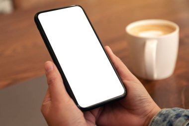 Masasında laptop ve kahve fincanı olan ve boş masaüstü ekranı olan siyah cep telefonunu tutan ellerin görüntüsü.