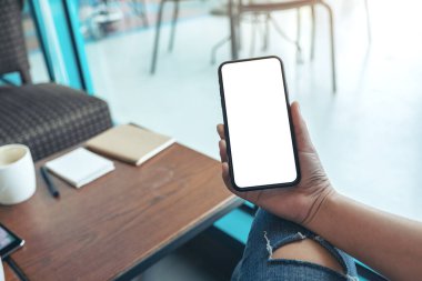 Kafedeki boş beyaz ekranlı siyah cep telefonlu bir kadının ellerinin görüntüsü.