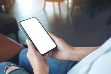 Kafedeki boş beyaz ekranlı siyah cep telefonlu bir kadının ellerinin görüntüsü.