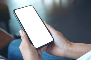 Kafedeki boş beyaz ekranlı siyah cep telefonlu bir kadının ellerinin görüntüsü.