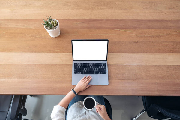 Top view mockup image of a woman using and touching on laptop touchpad with blank white desktop screen while drinking coffee on wooden table in office