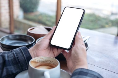 Masanın üzerinde kahve fincanı ve boş masaüstü ekranı olan siyah cep telefonunu tutan ellerin görüntüsü.