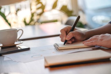 Bir kadının elleri deftere yazı yazıp iş bilgileri ve belgeler üzerinde çalışıyor.