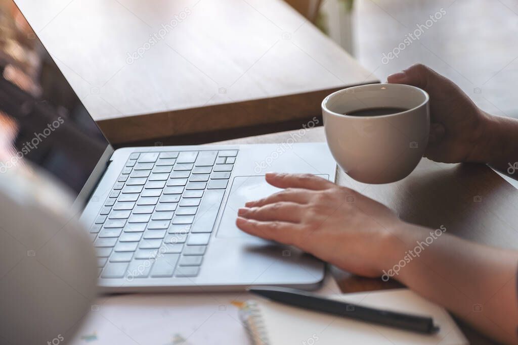Closeup image of a woman touching on laptop touchpad while drinking coffee and working in office