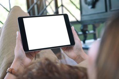 Oturma odasında rahatlamış bir şekilde uzanırken elinde beyaz ekranlı siyah tabletle yatay olarak duran bir kadının görüntüsü.