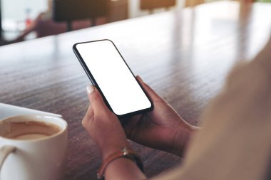 Masanın üzerinde kahve fincanı ve boş masaüstü ekranı olan siyah cep telefonunu tutan ellerin görüntüsü.