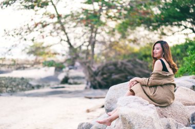 Güzel bir Asyalı kadın deniz kıyısındaki kayada oturmaktan zevk alır.