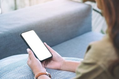 Oturma odasında rahat bir şekilde otururken elinde beyaz ekranlı siyah cep telefonu olan bir kadının görüntüsü.