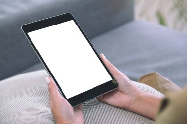 Oturma odasında rahat bir şekilde otururken elinde beyaz ekranlı siyah tablet bir kadının görüntüsü.