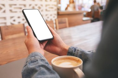 Masasında laptop ve kahve fincanı olan ve boş masaüstü ekranı olan siyah cep telefonunu tutan ellerin görüntüsü.