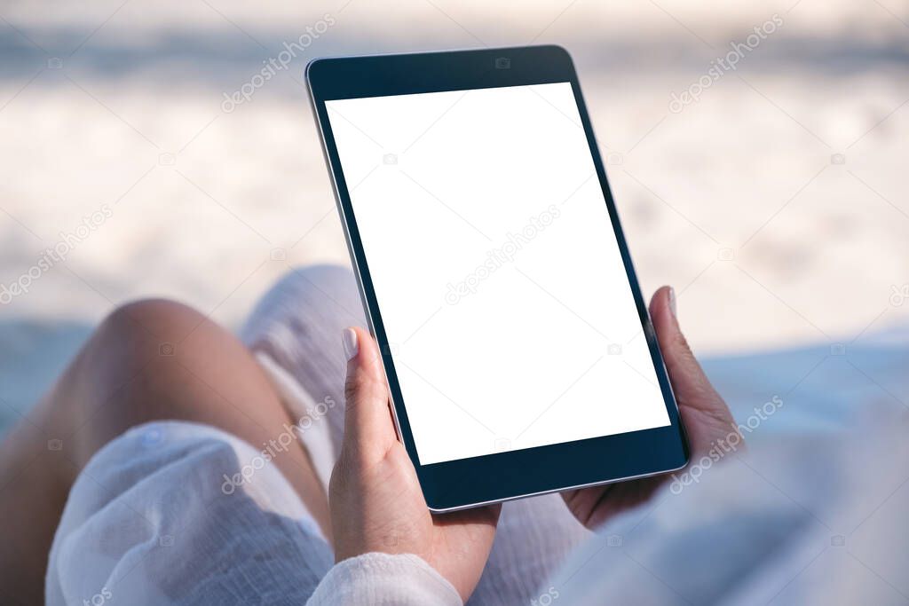 Mockup image of a woman holding a black tablet pc with blank desktop screen while laying down on beach chair on the beach